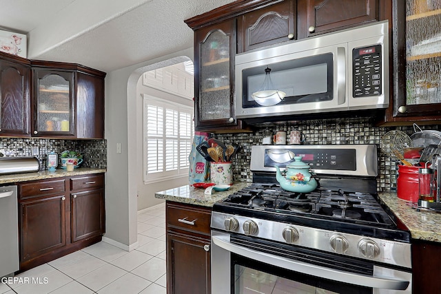 kitchen featuring light tile patterned floors, backsplash, stainless steel appliances, dark brown cabinetry, and light stone counters