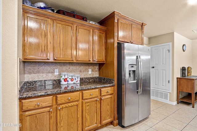 kitchen with stainless steel refrigerator with ice dispenser, dark stone countertops, light tile patterned flooring, and decorative backsplash