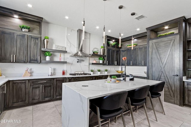 kitchen with hanging light fixtures, a kitchen island with sink, light stone counters, a barn door, and wall chimney exhaust hood