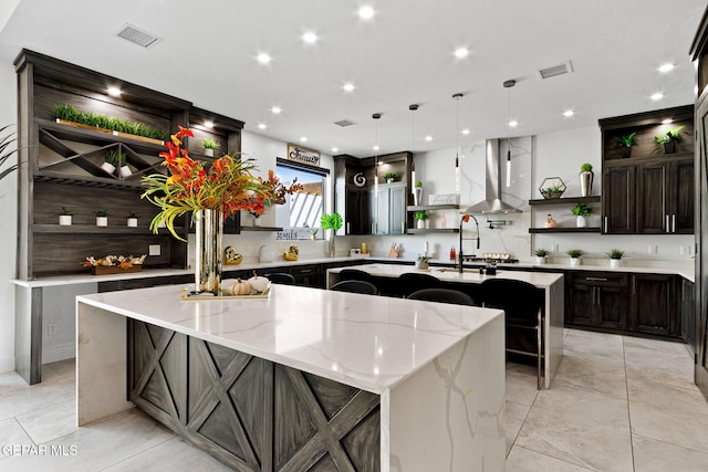 kitchen featuring wall chimney range hood, decorative light fixtures, light stone countertops, and a large island with sink