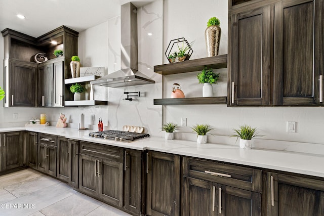 kitchen featuring range hood, stainless steel gas stovetop, decorative backsplash, dark brown cabinetry, and light stone counters