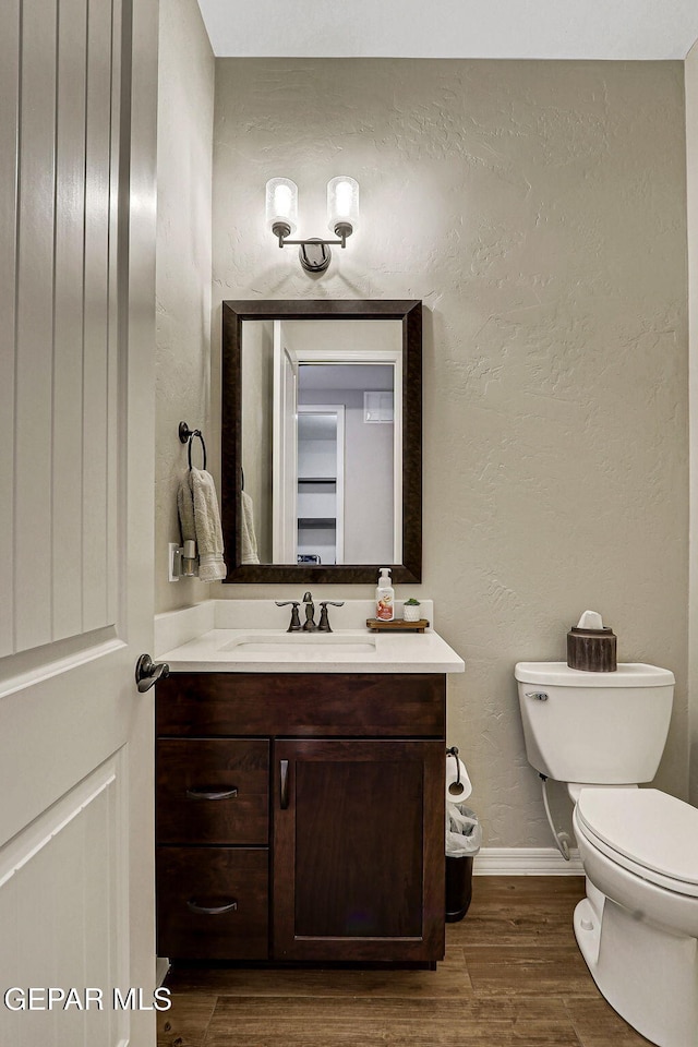 half bathroom featuring baseboards, toilet, wood finished floors, a textured wall, and vanity