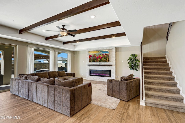 living area featuring stairway, beam ceiling, ceiling fan, light wood-type flooring, and a large fireplace