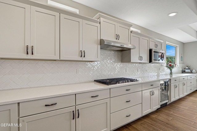 kitchen with beverage cooler, under cabinet range hood, backsplash, dark wood-style floors, and stainless steel appliances