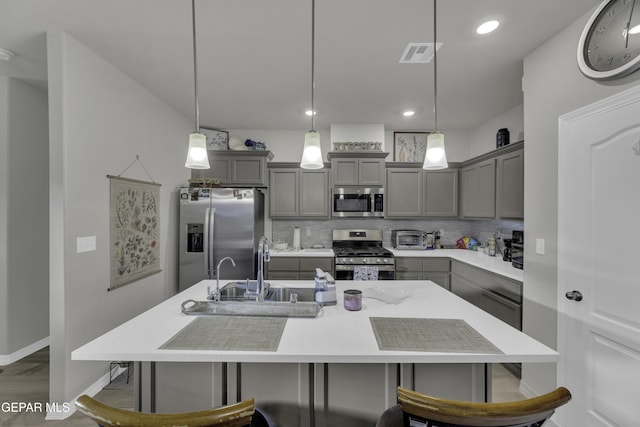 kitchen with gray cabinetry, an island with sink, pendant lighting, stainless steel appliances, and decorative backsplash