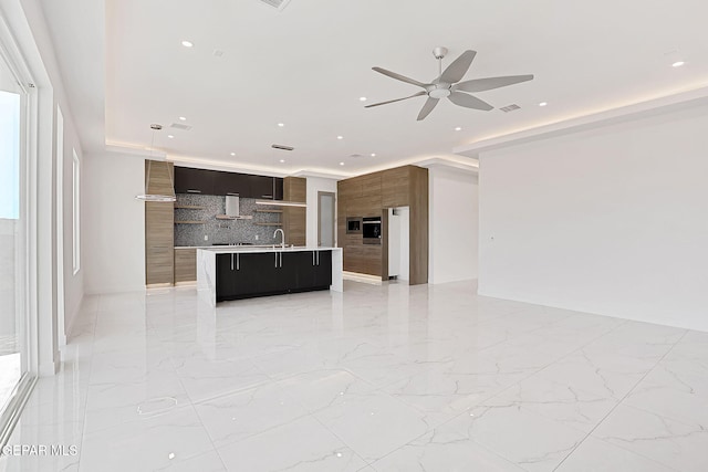 kitchen featuring wall chimney range hood, ceiling fan, a kitchen island with sink, backsplash, and a kitchen breakfast bar