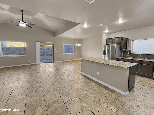 kitchen featuring a kitchen island, ceiling fan with notable chandelier, dark brown cabinetry, stainless steel refrigerator with ice dispenser, and light stone countertops