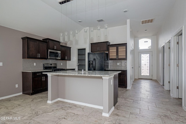 kitchen with appliances with stainless steel finishes, an island with sink, hanging light fixtures, light stone countertops, and dark brown cabinets