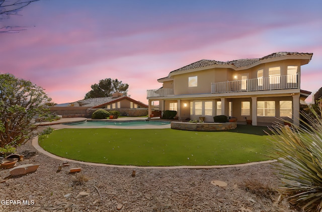 back of house at dusk with a balcony, a fenced in pool, a fenced backyard, and stucco siding