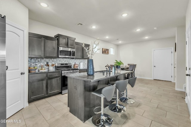 kitchen with sink, tasteful backsplash, dark stone countertops, appliances with stainless steel finishes, and a kitchen island with sink