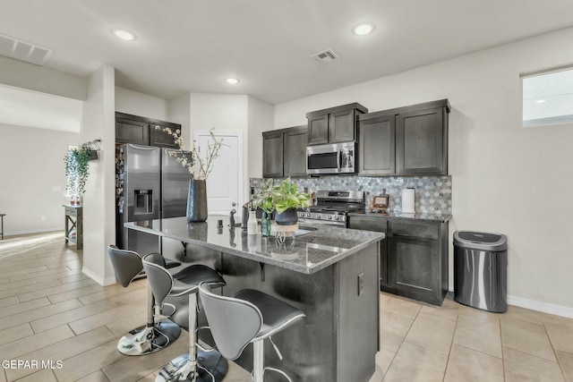 kitchen with dark brown cabinets, dark stone countertops, a center island with sink, stainless steel appliances, and backsplash