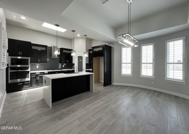 kitchen with appliances with stainless steel finishes, decorative light fixtures, an island with sink, sink, and wall chimney range hood