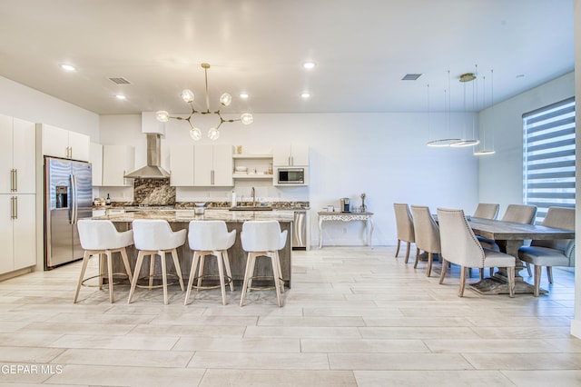 kitchen featuring pendant lighting, appliances with stainless steel finishes, an island with sink, white cabinets, and wall chimney exhaust hood