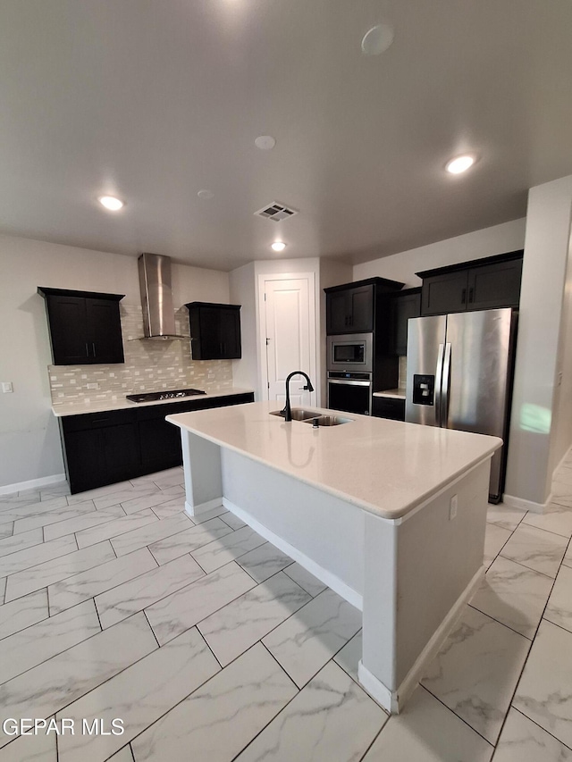 kitchen featuring wall chimney range hood, sink, appliances with stainless steel finishes, tasteful backsplash, and an island with sink