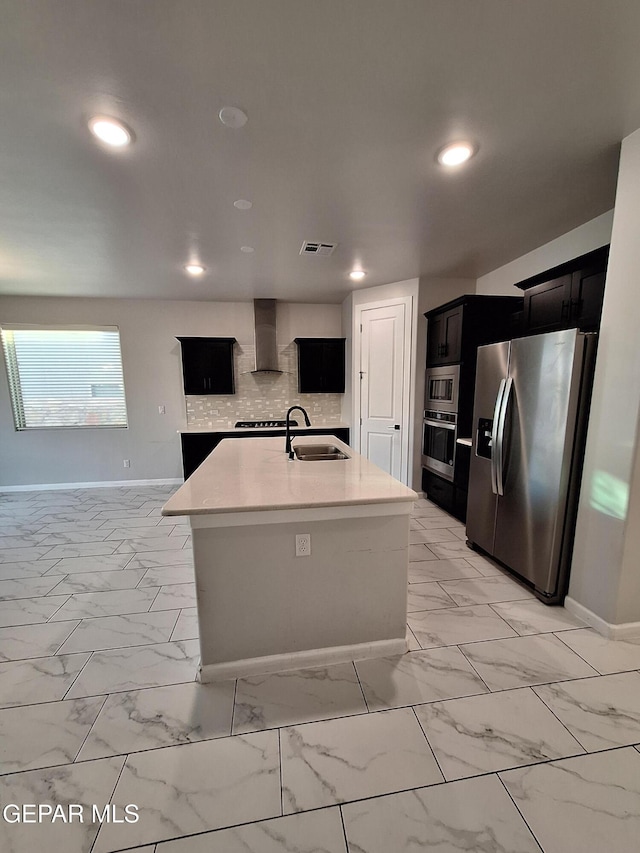 kitchen featuring wall chimney exhaust hood, sink, tasteful backsplash, a center island with sink, and appliances with stainless steel finishes