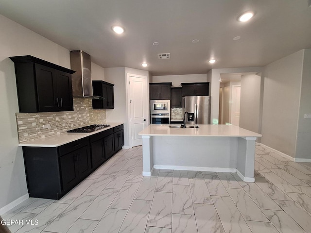 kitchen featuring sink, tasteful backsplash, a center island with sink, stainless steel appliances, and wall chimney range hood