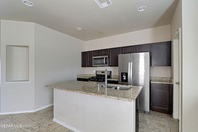 kitchen featuring sink, dark brown cabinets, stainless steel appliances, light stone countertops, and a center island with sink