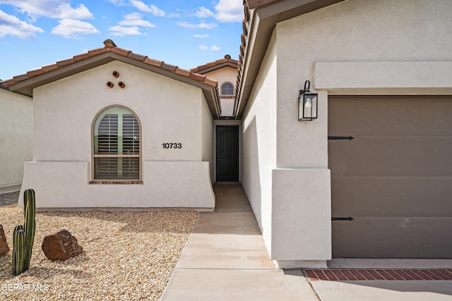 view of exterior entry with a garage, a tiled roof, and stucco siding