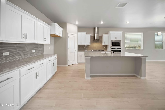 kitchen featuring appliances with stainless steel finishes, white cabinetry, light stone countertops, wall chimney exhaust hood, and light wood-type flooring