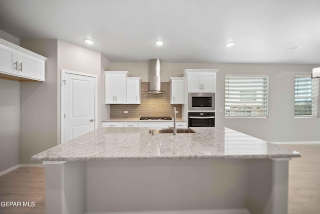 kitchen with white cabinetry, wall chimney range hood, light stone countertops, and a center island with sink