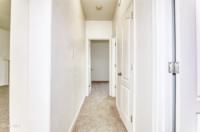 hallway with light carpet, a textured wall, a textured ceiling, and baseboards