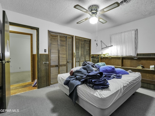 bedroom featuring ceiling fan, wooden walls, carpet floors, a textured ceiling, and multiple closets