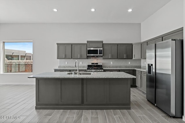 kitchen featuring sink, gray cabinets, appliances with stainless steel finishes, light stone counters, and a center island with sink