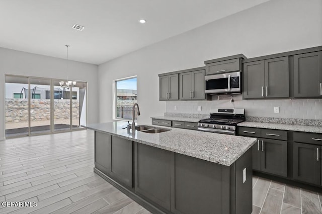 kitchen featuring appliances with stainless steel finishes, decorative light fixtures, an island with sink, sink, and light stone countertops