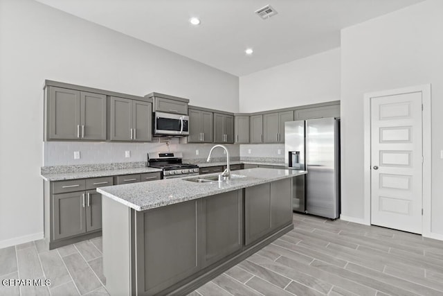 kitchen with sink, gray cabinets, stainless steel appliances, light stone countertops, and a center island with sink