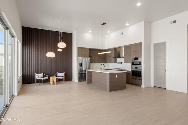 kitchen with hanging light fixtures, stainless steel appliances, a high ceiling, an island with sink, and light wood-type flooring