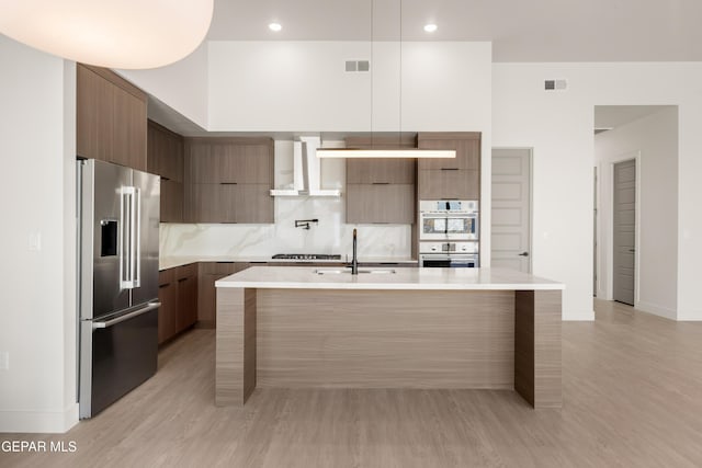 kitchen with wall chimney range hood, sink, stainless steel appliances, tasteful backsplash, and an island with sink