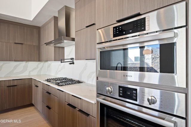 kitchen with appliances with stainless steel finishes, tasteful backsplash, wall chimney exhaust hood, light stone countertops, and light hardwood / wood-style flooring