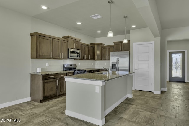 kitchen featuring appliances with stainless steel finishes, light stone counters, decorative light fixtures, a kitchen island with sink, and tasteful backsplash