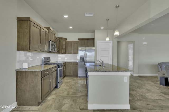 kitchen with stainless steel appliances, an island with sink, dark stone counters, decorative light fixtures, and sink