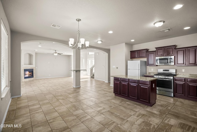 kitchen with a tiled fireplace, a kitchen island, light stone countertops, appliances with stainless steel finishes, and pendant lighting