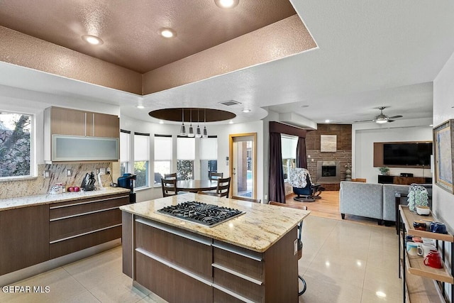 kitchen featuring a brick fireplace, light tile patterned floors, a center island, stainless steel gas stovetop, and tasteful backsplash