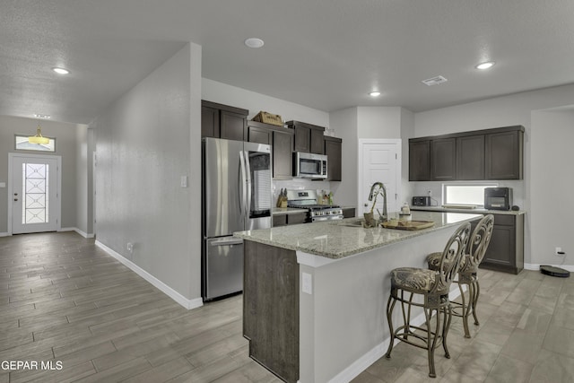 kitchen featuring an island with sink, sink, light stone counters, appliances with stainless steel finishes, and a breakfast bar area