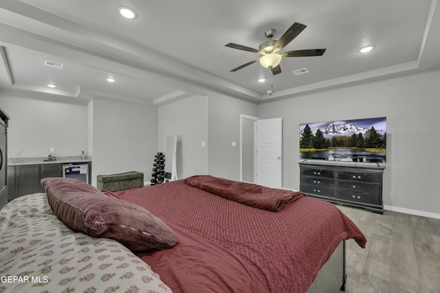 bedroom with indoor bar, light hardwood / wood-style flooring, wine cooler, a tray ceiling, and ceiling fan