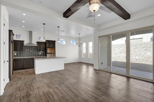 kitchen featuring a kitchen island with sink, beamed ceiling, wall chimney exhaust hood, and decorative light fixtures