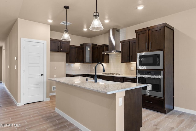 kitchen featuring a kitchen island with sink, visible vents, appliances with stainless steel finishes, light stone countertops, and wall chimney exhaust hood