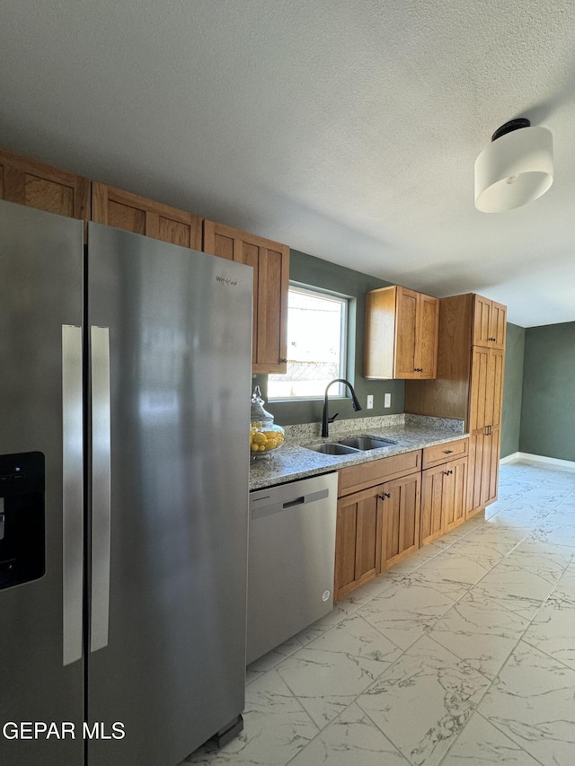 kitchen with a textured ceiling, stainless steel appliances, a sink, marble finish floor, and brown cabinets