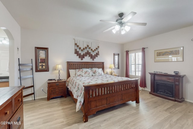 bedroom with arched walkways, a fireplace, a ceiling fan, baseboards, and light wood-style floors