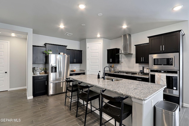 kitchen featuring a breakfast bar area, stainless steel appliances, visible vents, wall chimney range hood, and an island with sink