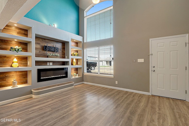 unfurnished living room featuring built in features, a high ceiling, a glass covered fireplace, wood finished floors, and baseboards