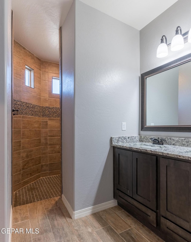 full bath featuring wood finish floors, tiled shower, vanity, and baseboards