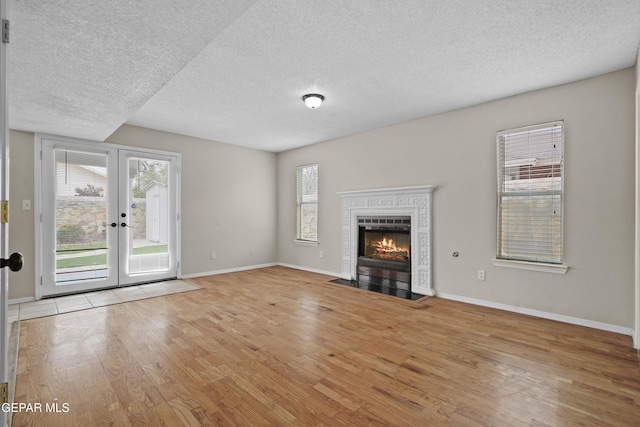 unfurnished living room featuring a textured ceiling, a fireplace with flush hearth, baseboards, light wood-style floors, and french doors