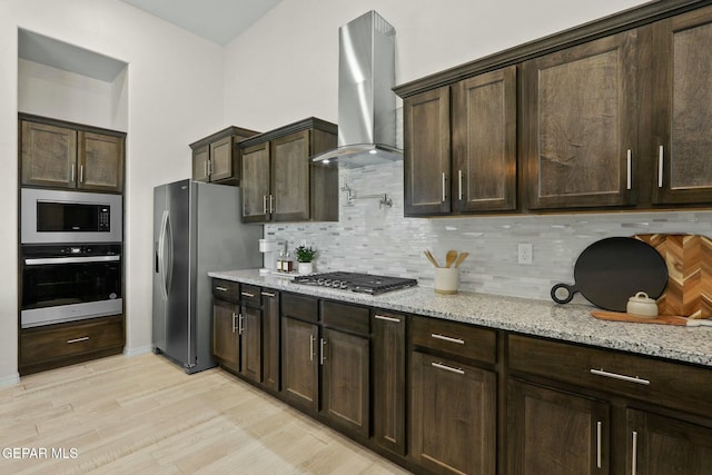 kitchen featuring stainless steel appliances, dark brown cabinets, wall chimney range hood, light stone countertops, and tasteful backsplash