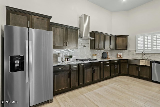 kitchen featuring light wood finished floors, wall chimney exhaust hood, appliances with stainless steel finishes, light stone countertops, and a sink