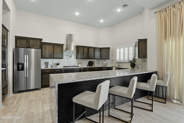 kitchen with light stone counters, a peninsula, exhaust hood, appliances with stainless steel finishes, and a kitchen bar