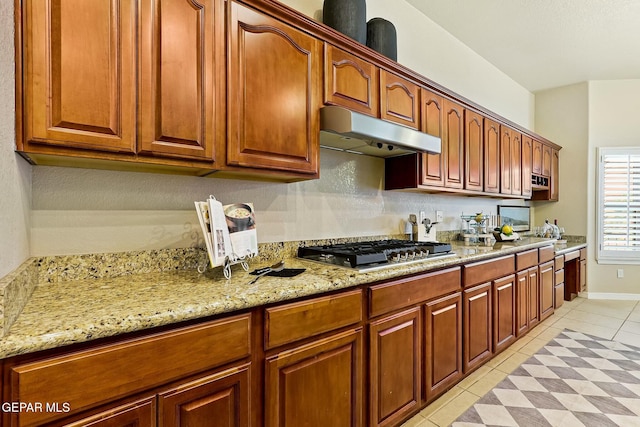 kitchen with brown cabinets, light stone countertops, under cabinet range hood, stainless steel gas cooktop, and light tile patterned flooring